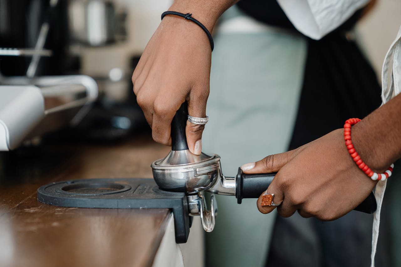Crop unrecognizable professional ethnic barista tempering ground coffee in portafilter using barista tools while working in professional coffeehouse